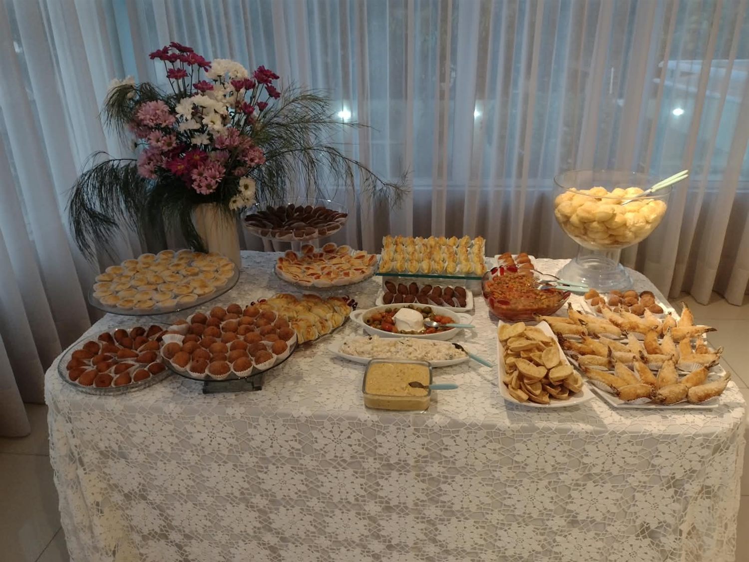 Mesa com toalha de renda, flores e salgados variados